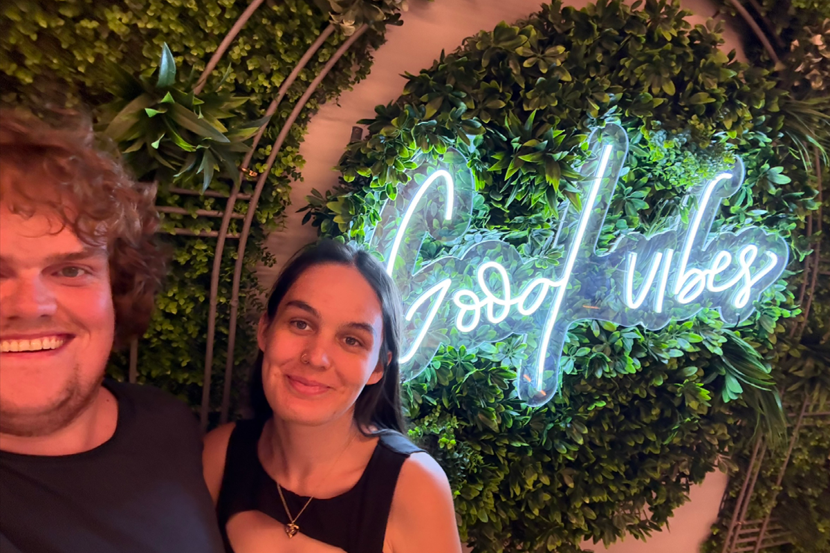 picture of two people smiling with a neon sign that says 'Good Vibes' behind them